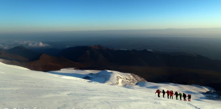 A team of seven climbers with their Russian guide in the lead ascending towards Lenz Rocks in the early morning. The quality of the light on the glaciers of Mount Elbrus just after sunrise is exceptional, due to the vast and icy slopes of the mountain lighting up instantly as soon as the sun hits.