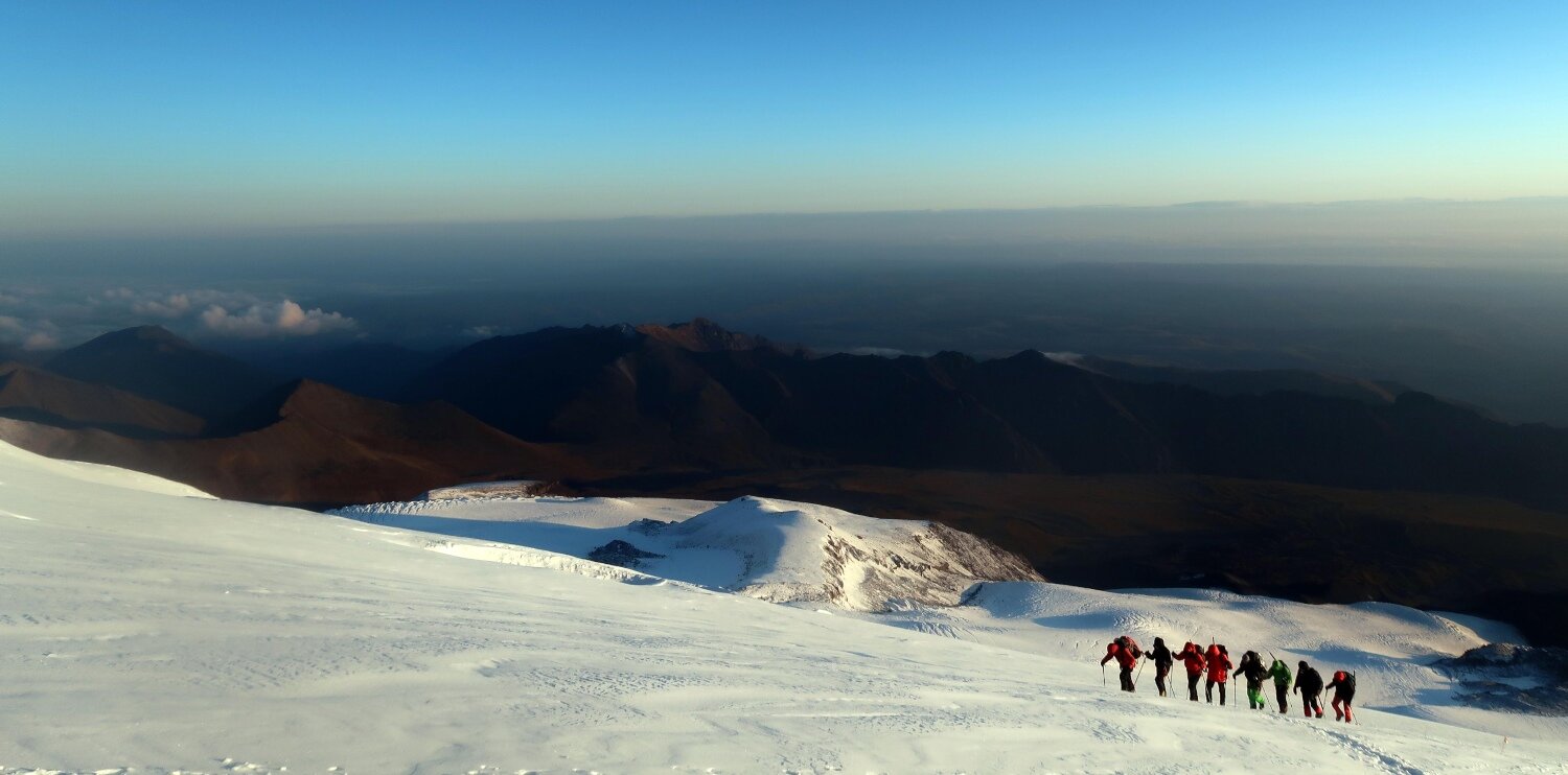 A team of seven climbers with their Russian guide in the lead ascending towards Lenz Rocks in the early morning. The quality of the light on the glaciers of Mount Elbrus just after sunrise is exceptional, due to the vast and icy slopes of the mountain lighting up instantly as soon as the sun hits.