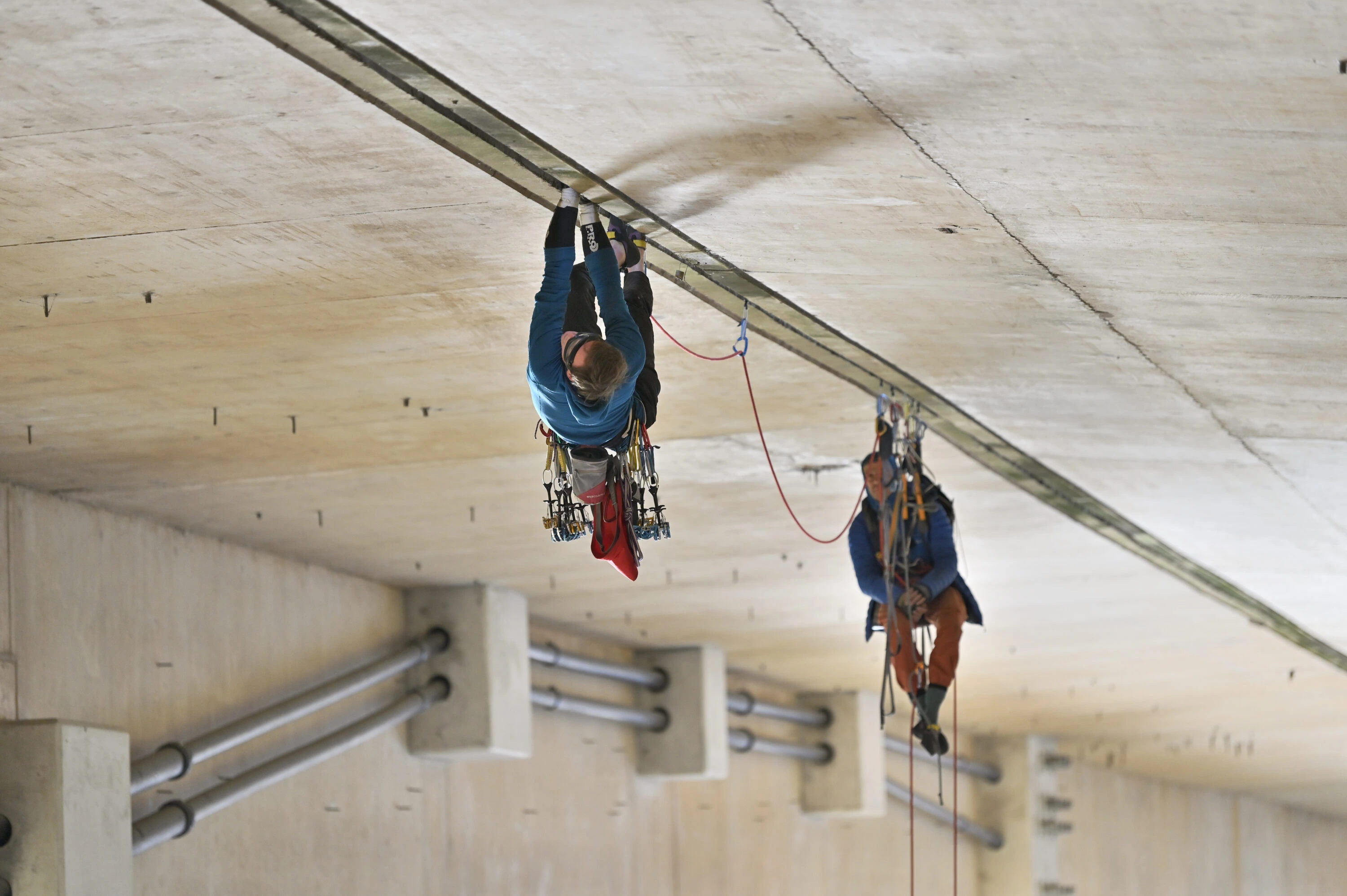 Two climbers climbing along a gap in the underside of a motorway bridge in the UK